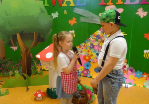 Dziewczynka trzyma w ręku mikrofon zwrócona w stronę chłopca recytuje wiersz. W tle dekoracja lasu.